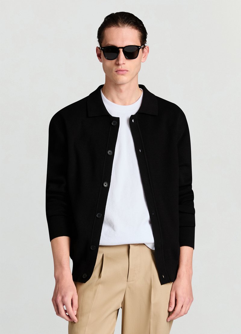 Cardigan en maille noire avec un col chemise, fermeture par boutons, et poignets côtelés, porté sur une chemise blanche, associé à un pantalon beige.