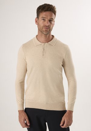 Homme portant un polo beige à manches longues avec trois boutons et un pantalon noir, se tenant devant un fond clair uni.