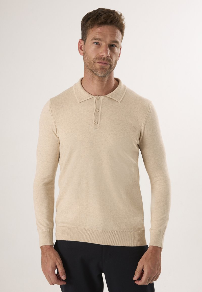 Homme portant un polo beige à manches longues avec trois boutons et un pantalon noir, se tenant devant un fond clair uni.