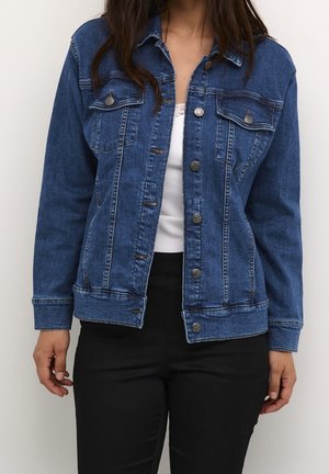 Femme portant une veste en denim bleue déboutonnée par-dessus un haut blanc et un pantalon noir, debout devant un fond clair uni.