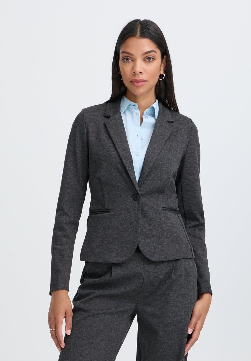 Femme aux longs cheveux foncés portant un ensemble blazer et pantalon gris foncé avec une chemise bleu clair boutonnée sur un fond uni.