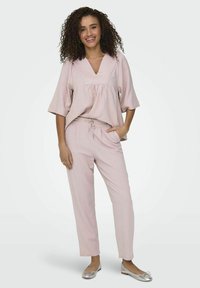Lichtroze, losvallend shirt met een V-hals en korte mouwen, gedragen met bijpassende broek met trekkoord; model draagt zilveren flats.