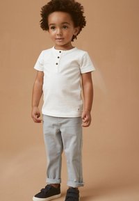 Hvid kortærmet henley skjorte med tre brune knapper, parret med lysegrå opsmøgede bukser og marineblå sneakers. Ensfarvet baggrund.