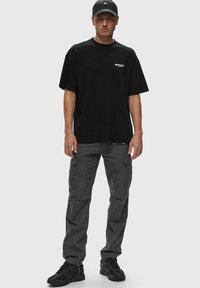 Camiseta negra con logo en el pecho, combinada con pantalones cargo grises con múltiples bolsillos y zapatillas negras. Diseño simple, estilo casual.