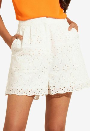Weiße Shorts aus Spitze mit gewelltem Saum, die mit ausgeschnittenen Mustern und seitlichen Taschen versehen sind, kombiniert mit einem knallorangefarbenen Oberteil.