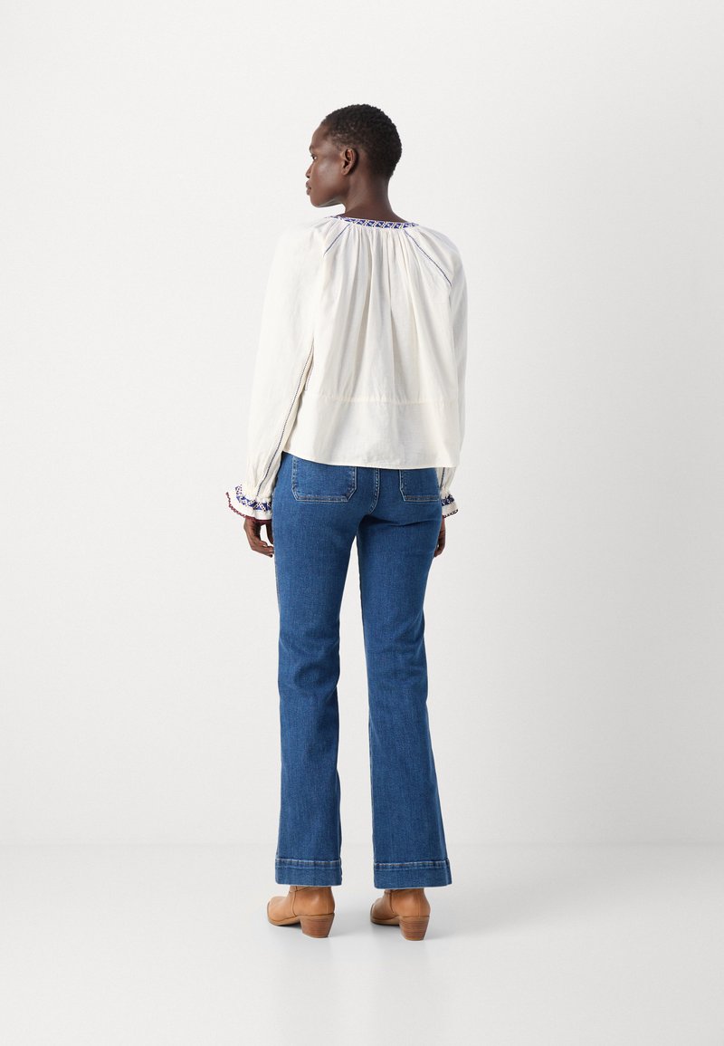 Chemisier blanc avec des détails froncés à l'arrière, rehaussé de broderies bleues. Associé à un jean évasé bleu et des chaussures à talons beiges.