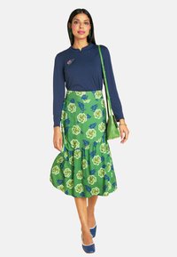 Eine Frau in einem navyfarbenen Langarmoberteil und einem grünen, blumigen Midikleid posiert, während sie eine kleine grüne Tasche hält, und stilvolle blaue Absätze ihr Outfit abrunden.