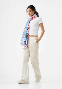 Écharpe multicolore avec des motifs, principalement bleue, présentant des designs géométriques et des accents, portée sur un haut blanc et un pantalon beige.