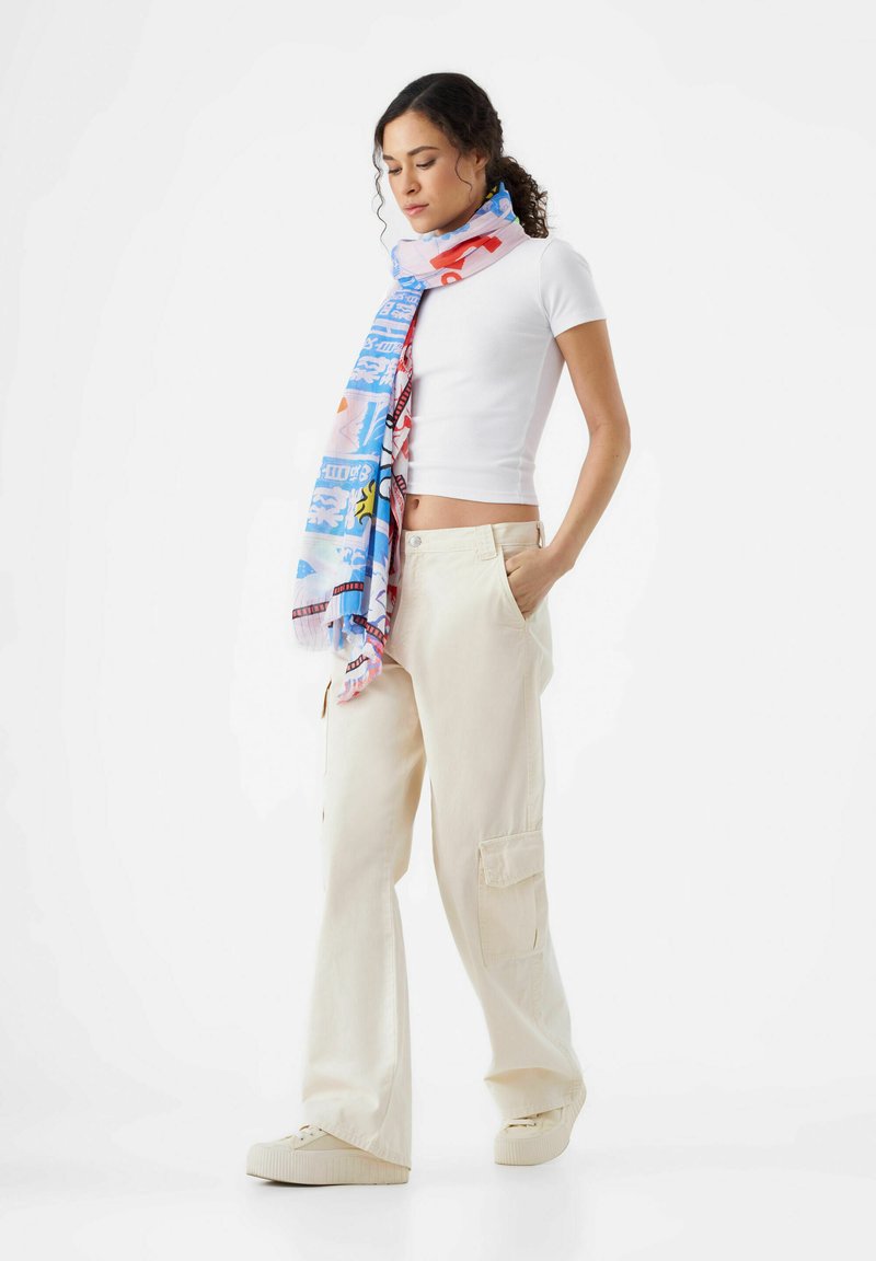 Écharpe multicolore avec des motifs, principalement bleue, présentant des designs géométriques et des accents, portée sur un haut blanc et un pantalon beige.