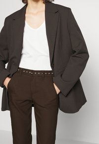 Blazer marron foncé sur une chemise blanche à col en V, associé à un pantalon marron avec une ceinture cloutée. Texture lisse et coupe ajustée.