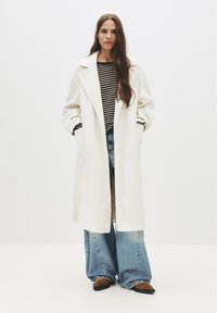 Biely dlhý kabát so širokými klopami, nosený cez čierno-biele pruhované tričko a modré široké nohavice. Hnedé špicaté balerínky dotvárajú celkový vzhľad.