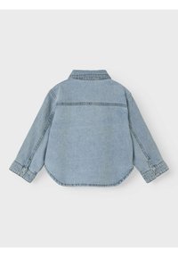 Camisa de mezclilla azul clara con collar puntiagudo, puños abotonados y dobladillo curvado. Presenta un detalle de costura en la yugo trasero.