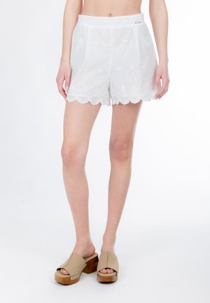 Pantalones cortos blancos de ojales con dobladillo festoneado, con motivos florales y una textura suave. Combinados con zuecos beige y una plataforma de madera.