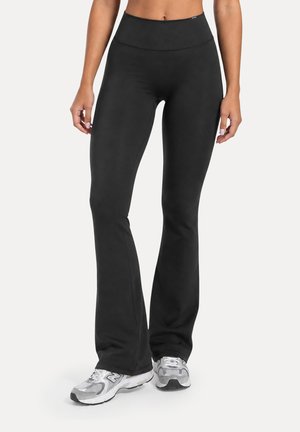 Mujer que lleva leggings negros acampanados de cintura alta combinados con zapatillas deportivas grises y blancas, de pie sobre un fondo liso.