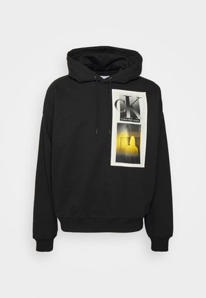 Sweat à capuche noir Calvin Klein avec un patch à l'avant affichant le texte "CK Calvin Klein Jeans" et un graphique de silhouette jaune.