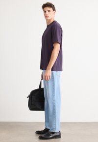 T-shirt en coton violet à manches courtes, associé à un jean en denim bleu clair. Le modèle tient un sac noir et porte des chaussures noires.
