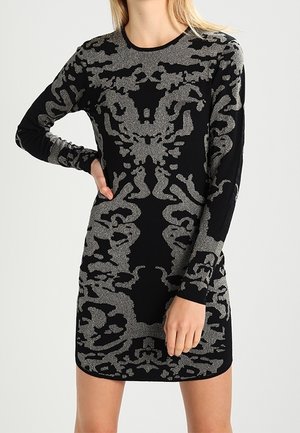 Femme portant une mini-robe noire à manches longues avec un motif floral abstrait gris complexe, debout avec une main sur la hanche.
