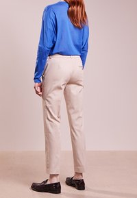 Kobieta z długimi włosami, ubrana w niebieską koszulę z długim rękawem, beżowe spędzone spodnie i czarne mokasyny, stoi na beżowej podłodze w tle neutralnej ściany.