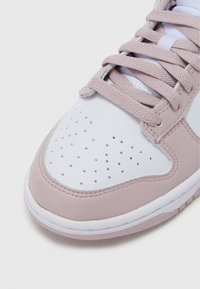 Urheilukenkä, jossa on valkoinen nahkainen päällinen vaaleanpunaisin yksityiskohdin, teksturoitu kärki, jossa on rei'itykset, ja litteät nauhat.