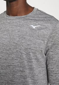Graues Langarm-Athletikshirt aus strukturiertem, meliertem Stoff mit weißem Logo auf der linken Brust. Glatte Halslinie und gestickte Akzente.