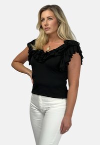 Czarny top bez rękawów z falbanami przy dekolcie, wykonany z miękkiego materiału, w połączeniu z białymi, dopasowanymi dżinsami.