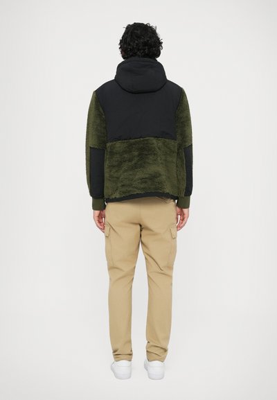 Jaqueta de fleece verde com a parte superior e capô pretos; combinada com calças cargo bege e sapatilhas brancas; mostrando uma vista traseira.