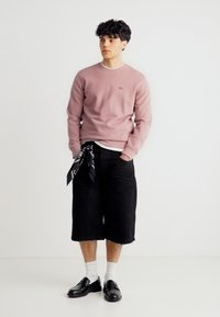 Νέος άνδρας που φοράει ροζ φούτερ, μαύρα φαρδιά σορτς μήκους μέχρι το γόνατο με μαύρο μπαντανα, λευκές κάλτσες και μαύρα μοκασίνια, στέκεται με τα χέρια στις τσέπες.