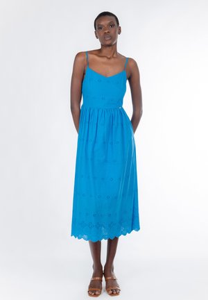 Blaues Baumwoll-Midikleid mit dünnen Trägern, mit Lochstickerei und gewelltem Saum. Modell steht in braunen Sandalen vor weißem Hintergrund.