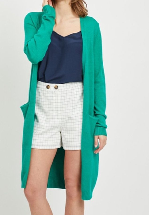 Cardigan long vert en matière douce, avec manches longues, poches latérales et poignets côtelés, porté sur un haut bleu marine et un short à carreaux.