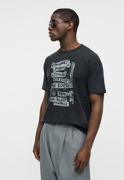 Άνδρας που φορά γυαλιά ηλίου, μαύρο γραφικό μπλουζάκι με φράσεις όπως «Ρομαντικά Ποιήματα» και «Νεαρό Πνεύμα», και γκρι πιετέ παντελόνι, στραμμένος προς τα αριστερά.