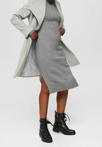 Robe grise en tricot avec une fente sur le côté, portée sous un manteau gris clair. Des bottines noires à lacets complètent la tenue.
