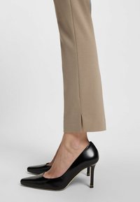 Zwarte lakleren hoge hakken met een spitse neus en slanke hak, gecombineerd met aansluitende beige broeken met een zijsplit aan de zoom.