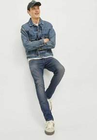 Chaqueta de denim en azul claro, con cuello clásico, dos bolsillos en el pecho. Vaqueros azules, ajustados. Zapatillas beige claras con cordones blancos y detalles grises.