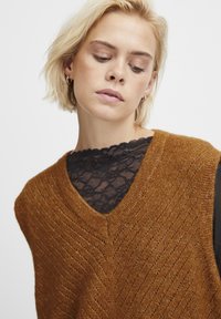 Bronzefarbenes, gestricktes Top mit tiefem V-Ausschnitt und strukturiertem Zickzackmuster, über einem schwarzen Spitzentop getragen. Kurzärmliges und leichtes Design.