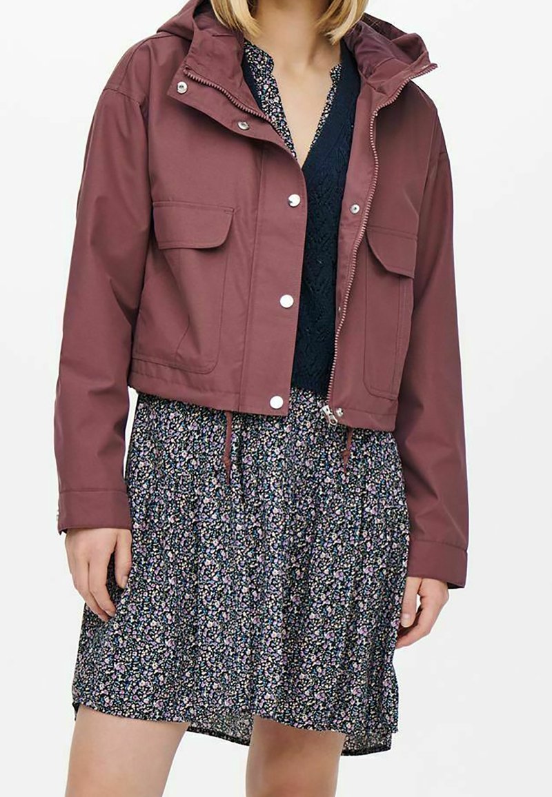 Przycięta burgundowa kurtka z kapturem, z srebrnymi napami, posiadająca dwie przednie kieszenie, noszona na kwiecistej sukience. Gładki materiał, casualowy design.
