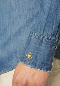 Cuff di camicia di denim con logo ricamato "LB" giallo e frecce direzionali, indossato sopra pantaloni chiari.