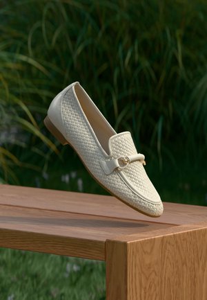 Mocassins tissés crème avec un accent en cuir lisse, semelle plate et détail en métal, posés sur une surface en bois contre un fond vert.