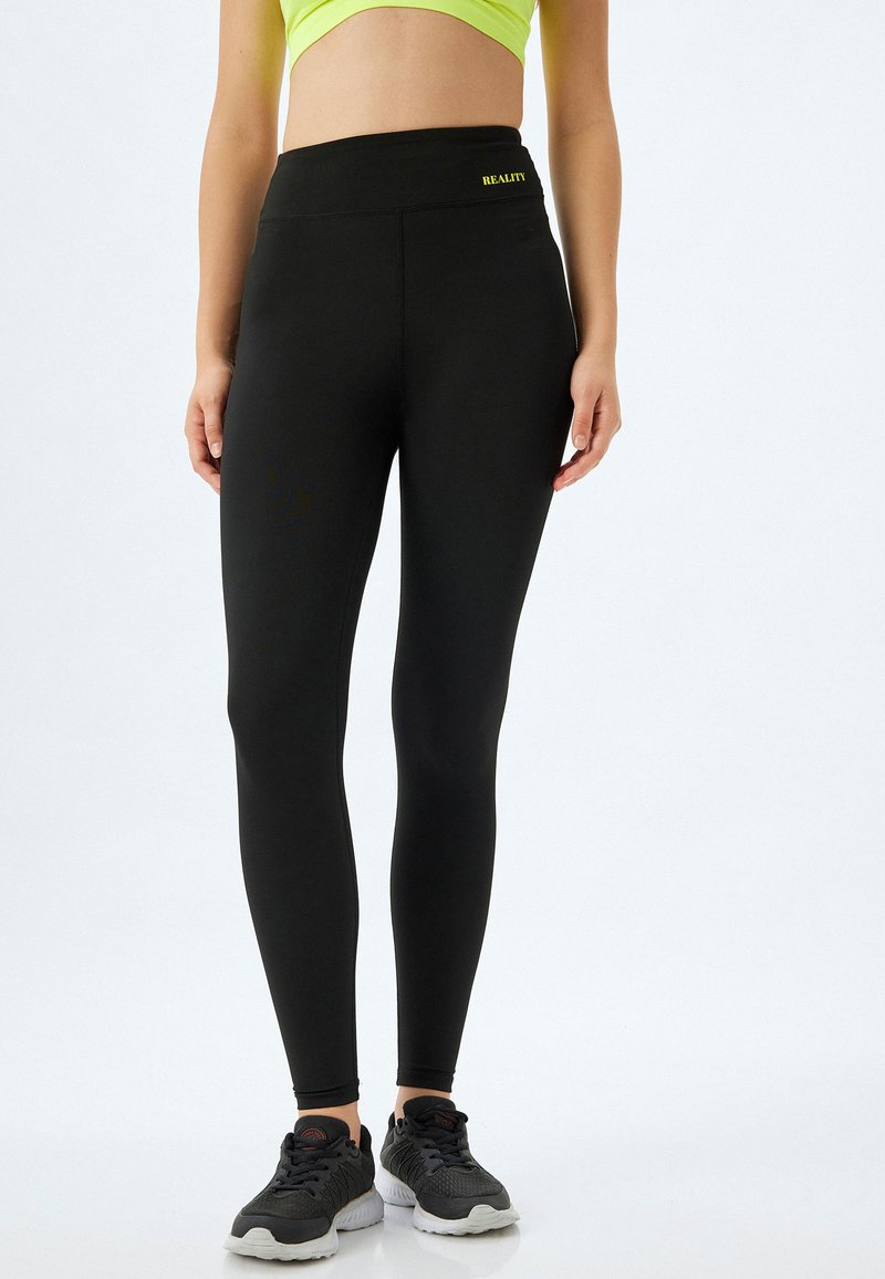 Zwarte leggings van rekbare stof, met een hoge tailleband en een kleine gele tekst "REALITY". Ideaal voor sportieve kleding.
