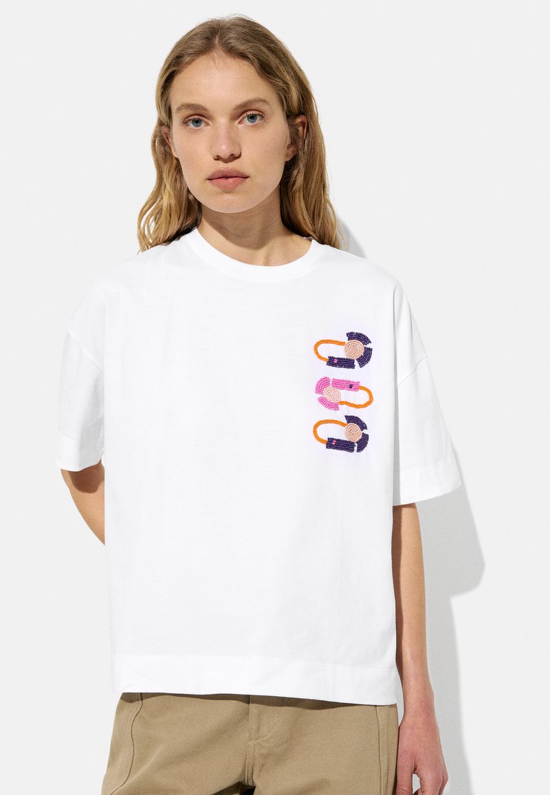 Biała bawełniana koszulka z okrągłym dekoltem i krótkimi rękawami. Na przodzie znajduje się kolorowy haftowany wzór graficzny.