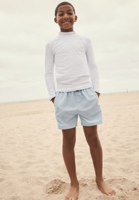 Maglia protettiva bianca a maniche lunghe e pantaloncini da bagno azzurri chiaro con vita elastica, in piedi su una spiaggia sabbiosa.