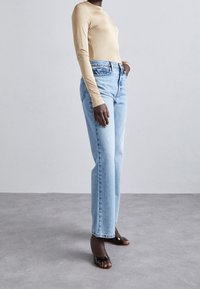 Beiges Langarmtop kombiniert mit hellblauen geraden Jeans mit hoher Taille und schwarzen offenen Blockabsätzen. Glatte Stoffqualität und lässiger Schnitt.