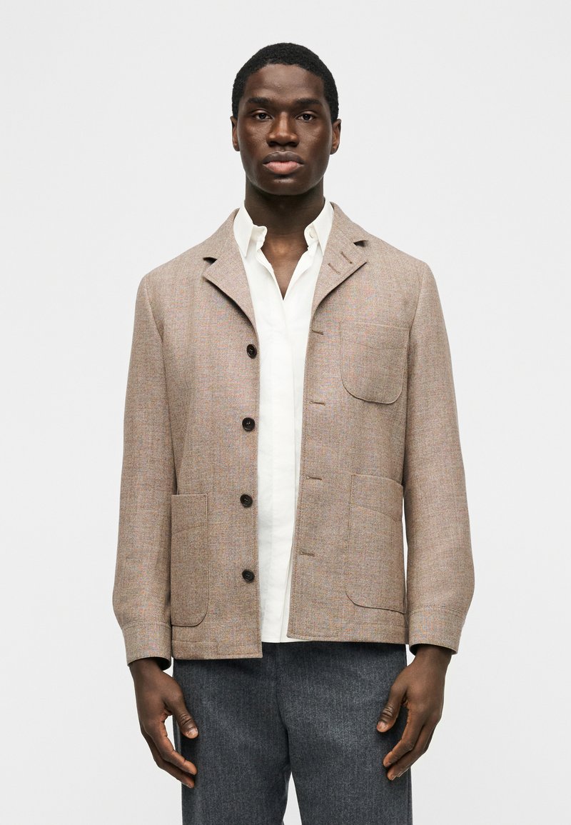 Άνδρας που φοράει μπεζ σακάκι με κουμπιά και τρε ις μπροστινές τσέπες πάνω από λευκό πουκάμισο και γκρι παντελόνι, στέκεται μπροστά σε απλό φόντο.