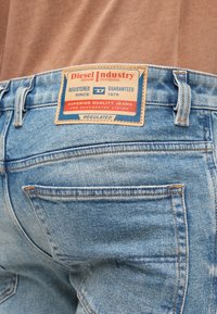 Vaqueros de mezclilla azul con un parche de cuero marrón que lleva texto en rojo y azul. La tela tiene textura y cuenta con bolsillos traseros estándar.