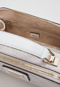 Sac à main Guess beige et blanc avec fermeture éclair dorée, anse blanche et doublure intérieure beige comprenant une poche zippée et une étiquette logo Guess.