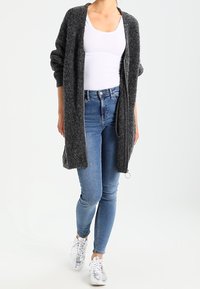 Lång, öppen cardigan i mörkgrått stickat material, bärs över en vit topp och kombineras med blå jeans och silverfärgade sneakers. Avslappnad design.