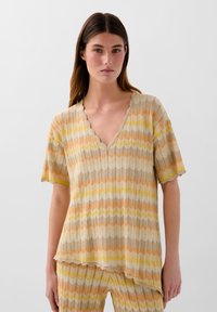 Knit top featuring a scalloped neckline, short sleeves, and a zigzag pattern in beige, orange, and yellow. Lightweight and textured fabric.