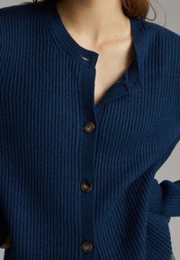 Granatowy żakiet prążkowany z dekoltem w kształcie litery V i pięcioma brązowymi guzikami, charakteryzujący się teksturowanym materiałem oraz detalem z otwartym splotem w okolicy kołnierza.