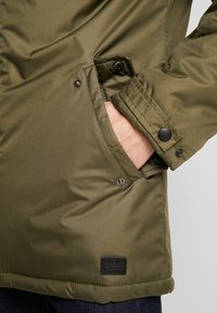 Veste matelassée vert olive à texture lisse, avec une poche latérale à fermeture par bouton-pression et une étiquette de marque noire sur l'ourlet.