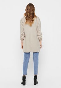Kvinna med vågigt hår klädd i en beige lång kappa, blå jeans med fransade fållar och svarta klackade anklarstövlar, stående mot en vit bakgrund.