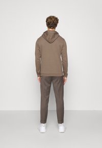 Hombre de pie, de espaldas, vestido con sudadera marrón, pantalones a juego y zapatillas blancas, frente a un fondo claro y liso.